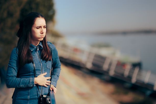 female tourist with long brown hair in a blue denim jacket with camera hanging from strap around her neck; boat dock in the background; the woman’s hand on her stomach and cheeks filled with air to show an ill feeling 