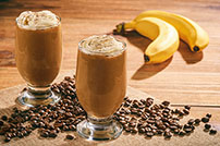 two glass mugs hold banana mocha smoothies with two bananas and coffee beans scattered on wooden board 623347650