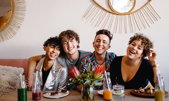 A group of four young, gender-fluid adults sitting on the couch with their arms around each other posing for the camera.