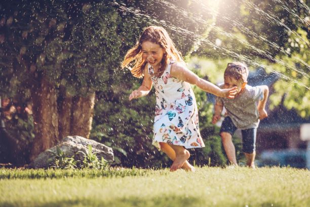 Girl and boy laughing while running through sprinkler on warm summer day 