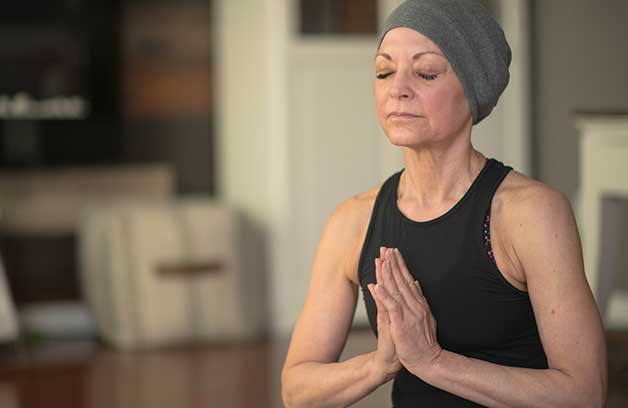 Senior woman with cancer, meditating while seated on the floor. She's wearing a gray head scarf and a black leotard.