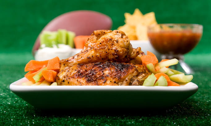 Slow cooker BBQ wings on a plate with carrots and celery with a football and bowl of chips in salsa in the background.