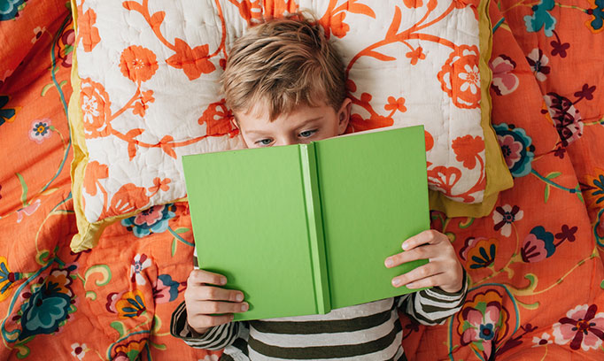 Little boy laying in bed struggling to read a book because he needs glasses