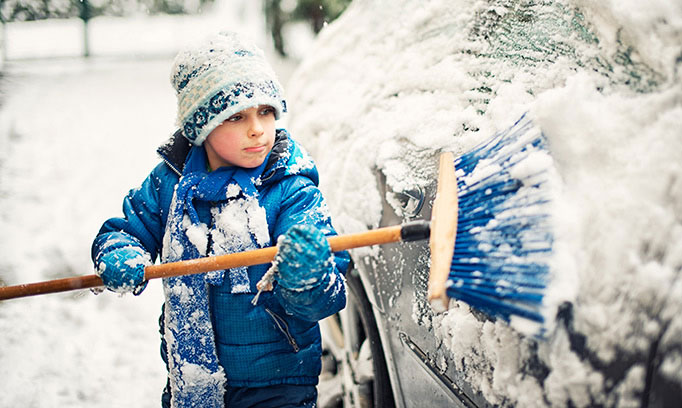 Snow-covered boy brushes snow off a car