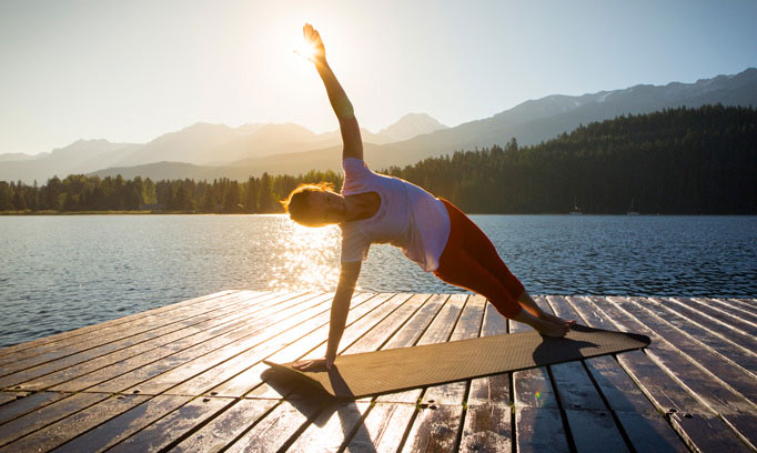 Woman holds yoga pose near mountain lake, a practice that may help with symptoms of multiple sclerosis (MS).. 