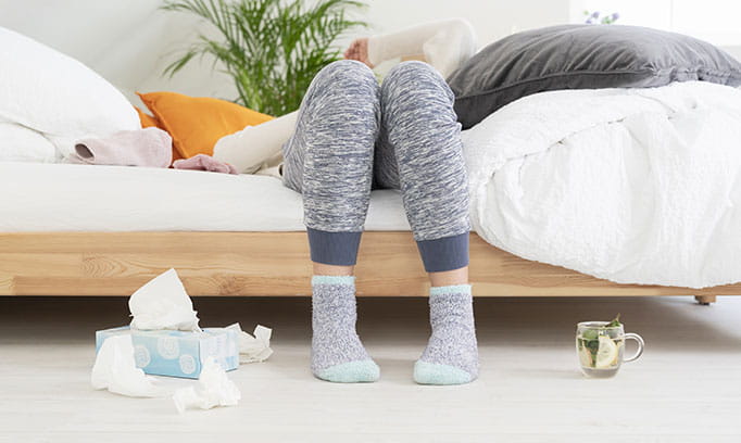Woman sitting on a bed and fallen backwards, surrounded by tissue boxes, used tissues and hot tea.