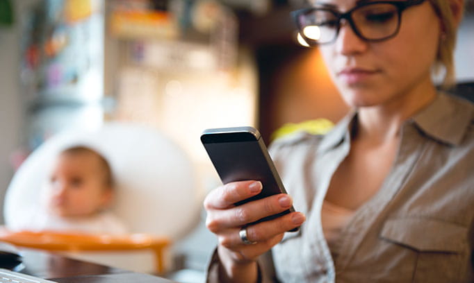 Young mom distracted by phone with baby in the background
