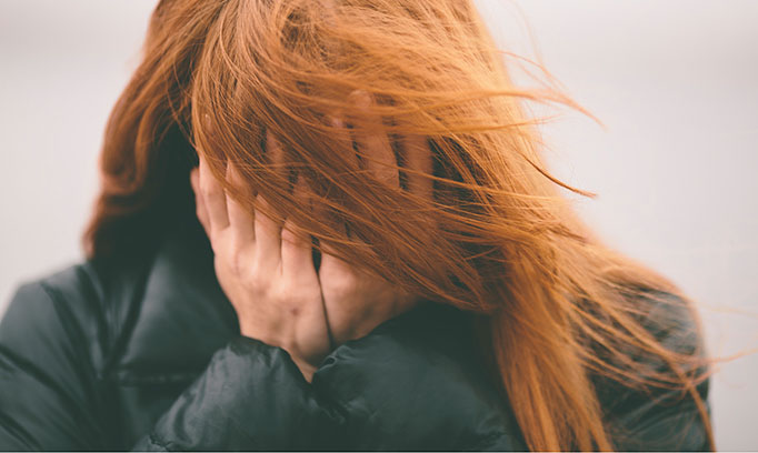 Woman's face is covered by her long red hair and her hands as though unable to face a stressful situation