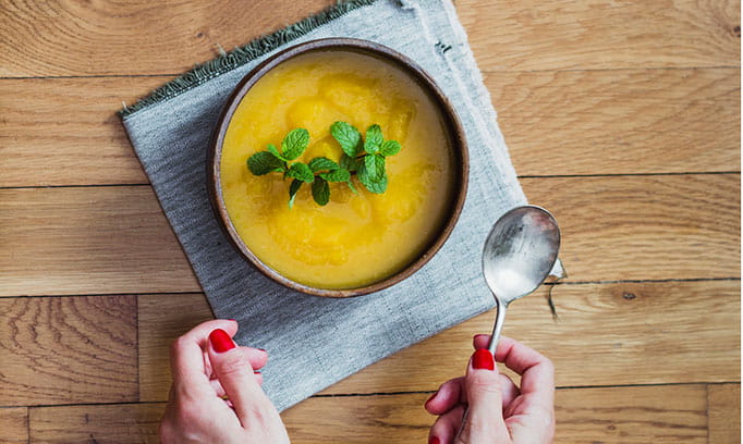 Bird's eye view of a bowl of pumpkin mushroom soup with a pair of hands reaching to pick it up.