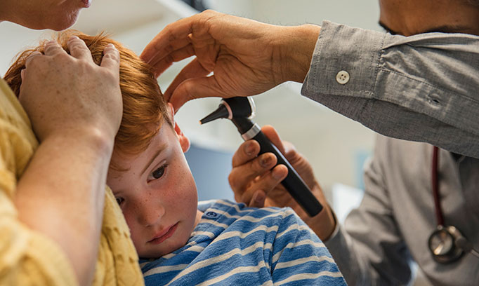young boy with symptoms of ear infection being held by mom has his ear examined by a doctor