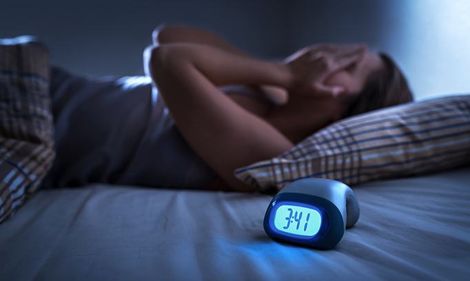 Pictured is a woman lying in bed with hands over her face. A digital clock reads 3:41 a.m.