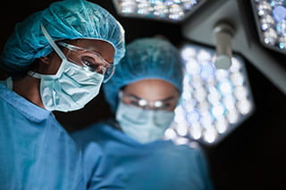 Surgeons in gowns, masks, caps, and protective eyewear operate under lights.