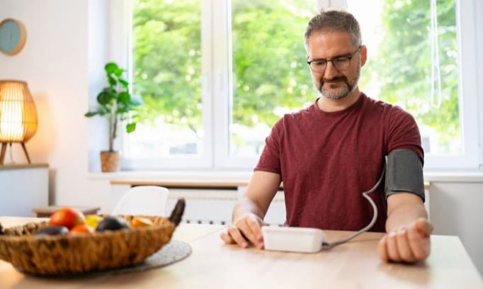 Man seated at a table wearing a blood pressure cuff and viewing a home BP monitor.
