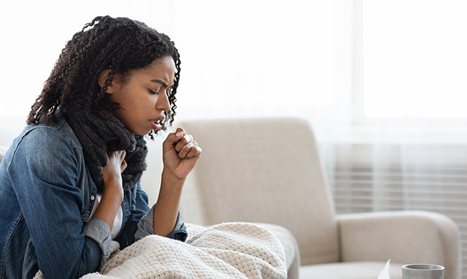 Young black woman with bronchitis coughs into her hand as she sits on white couch with white blanket
