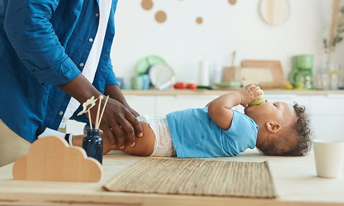 Black parent in denim jacket, shown from shoulders to midthigh, is changing the diaper of his newborn baby who is wearing a blue Tshirt and diaper