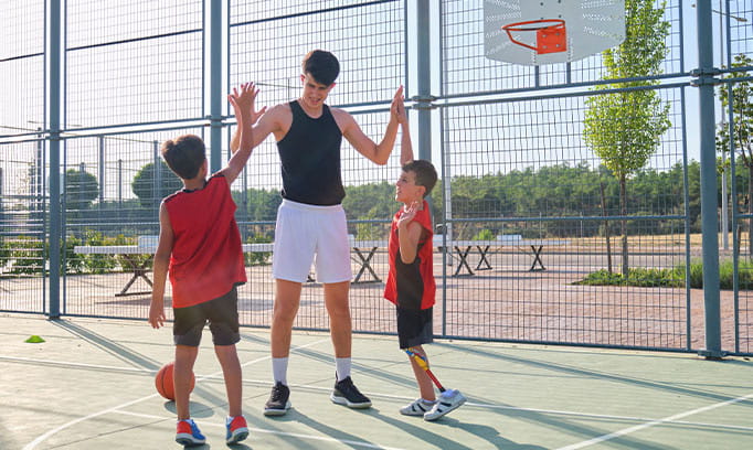2207 HSG Kids and Exercise a teen and two younger boys in shorts and tank tops give high fives on basketball court