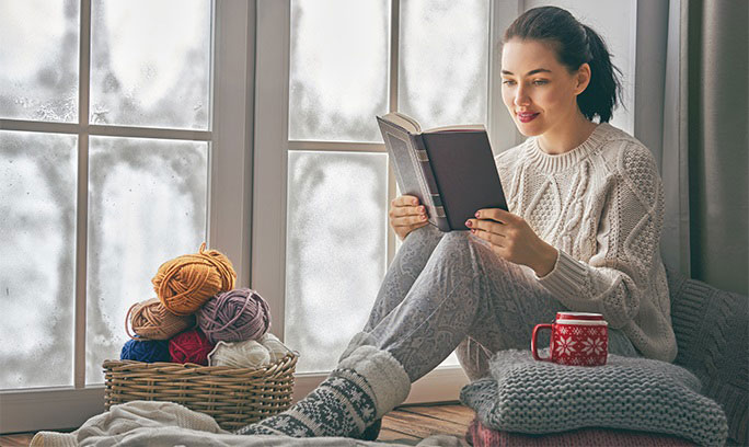 woman woman in cozy winter clothing sits in window nook reading and drinking a mug of hot tea