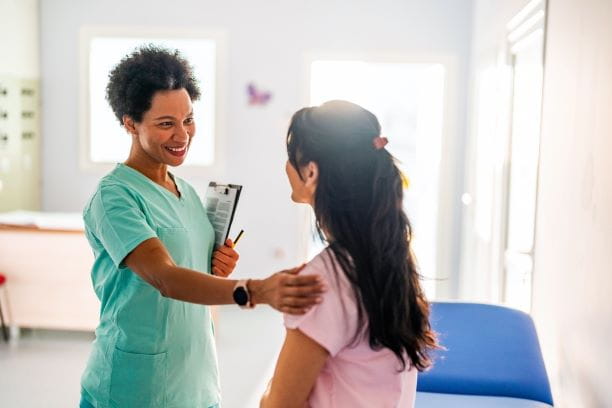 female doctor standing in front of seated female patient with a hand extended and resting on the patient’s shoulder in an exam setting