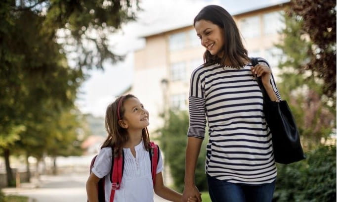 mother holding hand of daughter wearing backpack in front of school building
