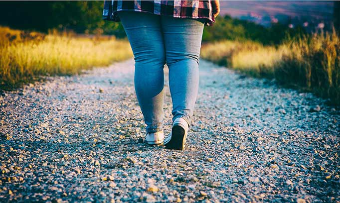 Hip down image of a woman walking down a straight hiking path surrounded by wild grass.