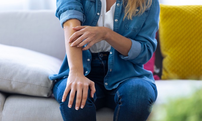 Woman siting on a couch itching her arm wondering what type of dermatitis she has.