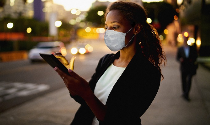 Black woman wearing a face mask checks her cell phone outdoors in city at dusk 682x408