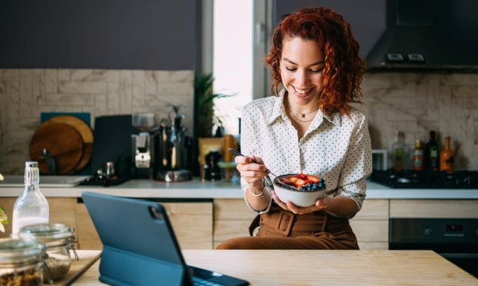 young woman in kitchen eating foods that relieve stress: bowl of oatmeal with fresh berries