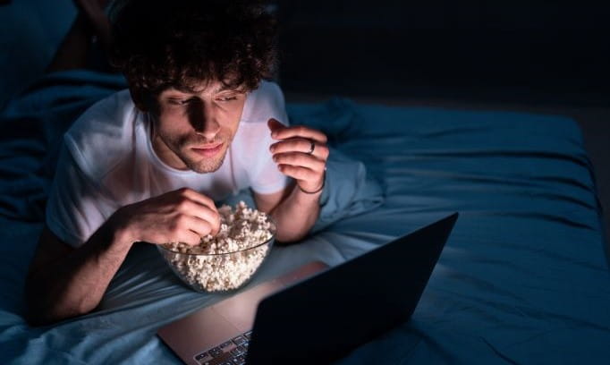 guy going goblin mode eating popcorn in dark and watching laptop