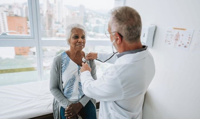 male doctor using stethoscope to listen to heartbeat of elderly woman in an exam room
