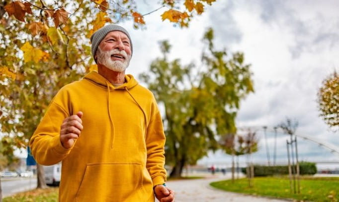 older man with short white beard wearing yellow hoodie and gray hat while walking on a sidewalk in the fall