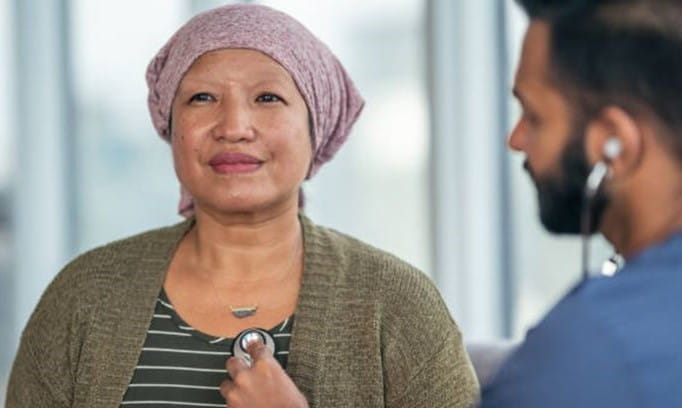 male medical professional using a stethoscope to check the heart of a female patient wearing a bandana to hide hair loss from chemotherapy treatment