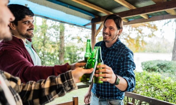 three men standing under a covered deck in a summer scene clinking green beer bottles together