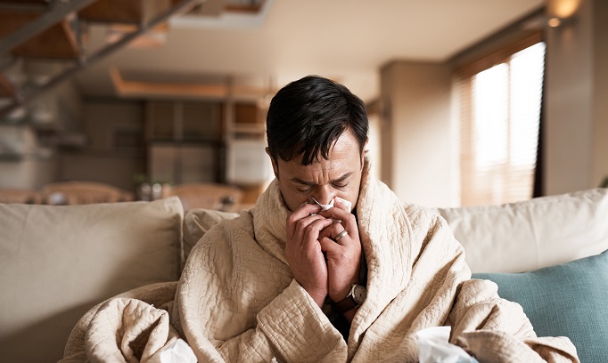 Man sitting on the sofa wrapped in a blanket, blowing his nose into a tissue because he has the flu