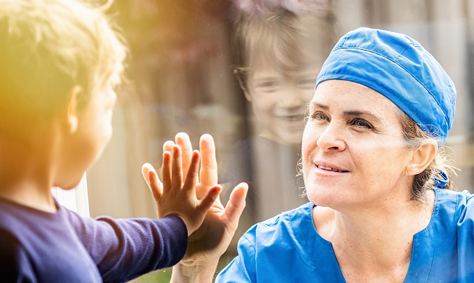 woman health care worker in scrubs smiles at child and they touch hands on window between them