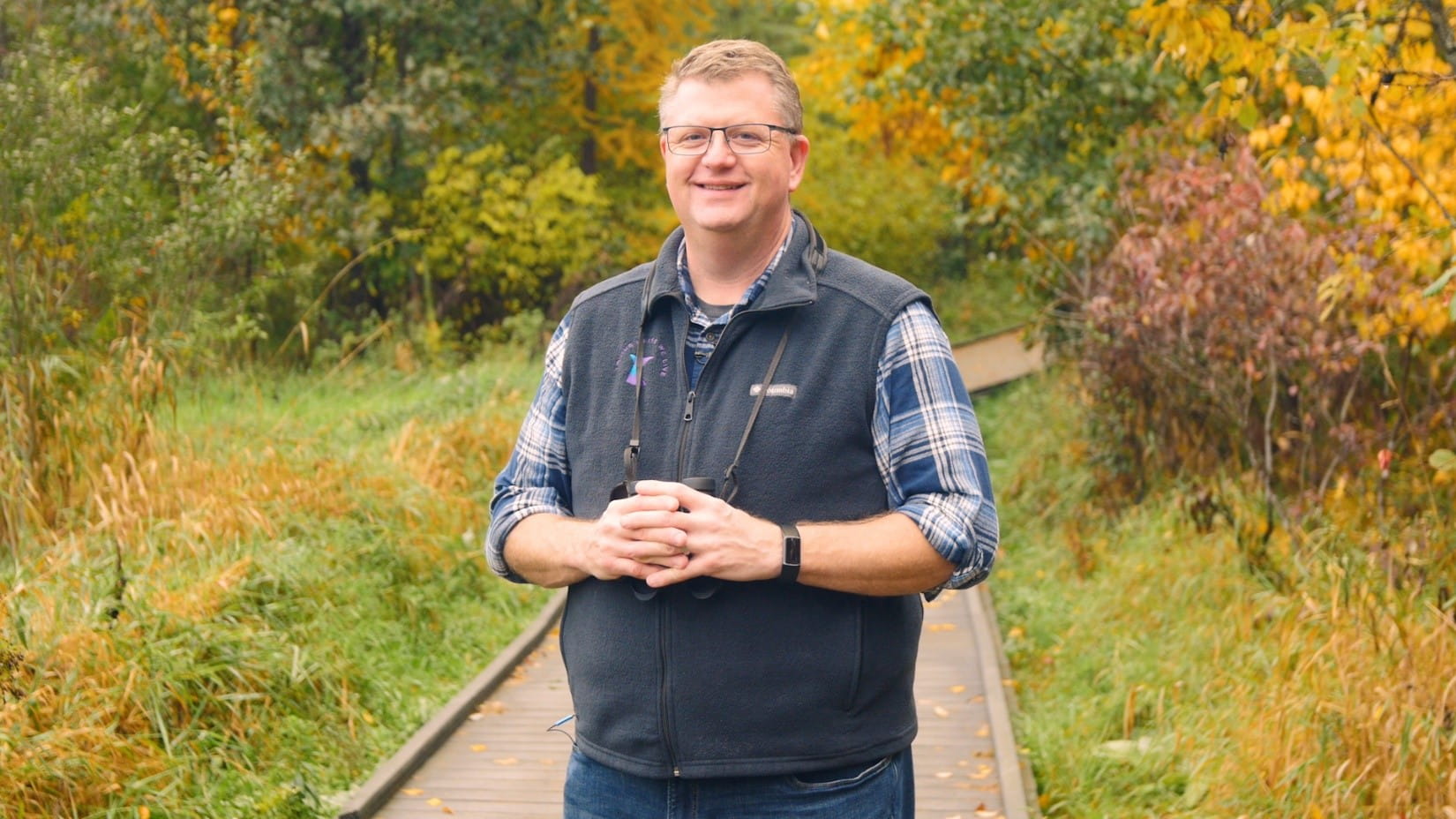a man in glasses, wearing a plaid shirt and vest, smiling at the camera while holding birdwatching binoculars hung around his neck with a thin strap