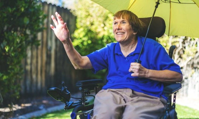 smiling woman with multiple sclerosis seated in a wheelchair waving right hand while holding yellow umbrella with left hand