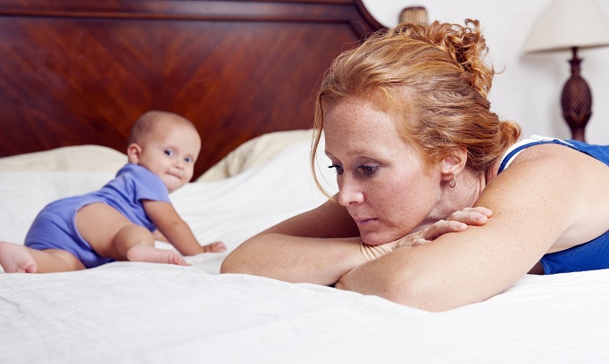 woman struggling with postpartum depression shown with baby who is crawling on bed