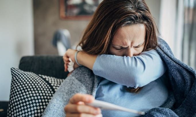 woman with respiratory virus coughing into elbow and holding thermometer