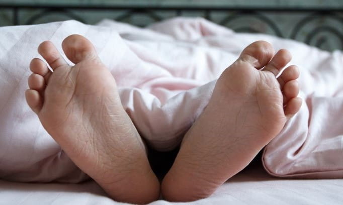 underside of an adult person’s feet sticking out from under a white blanket while lying in a bed 