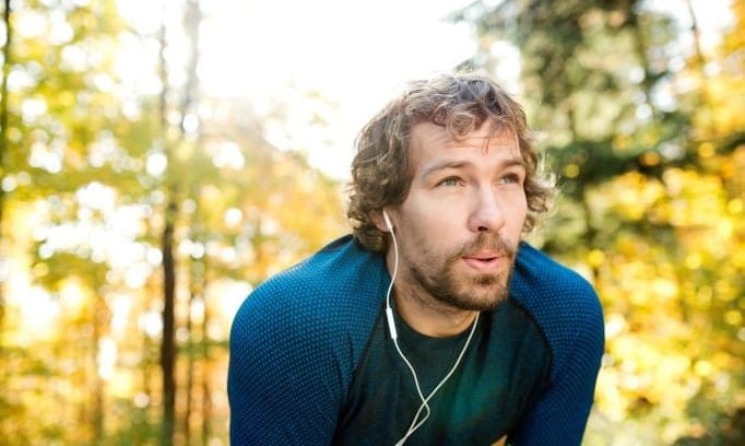 Young male exhaling while wearing ear buds crouching with hands on knees in an autumn woodland scene
