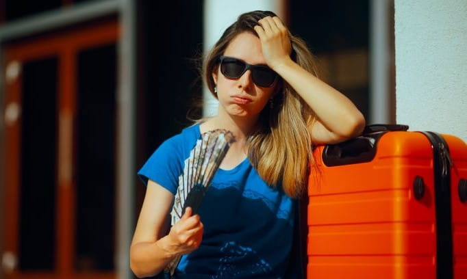 woman wearing sunglasses and waving a fan while sitting on a curb next to a suitcase