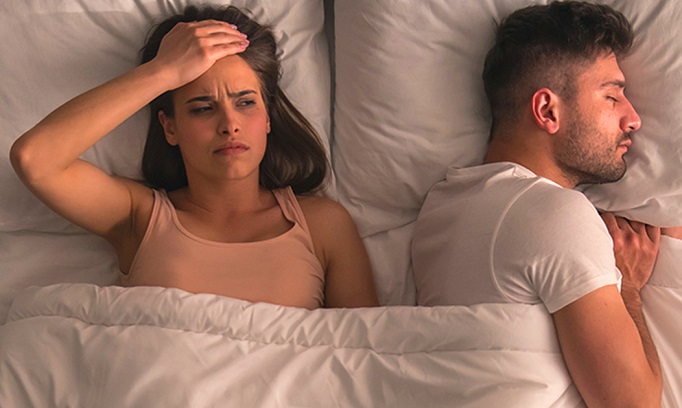 man and woman lie next to each other in bed woman awake with right hand on forehead because man on his left side sleeping may be snoring