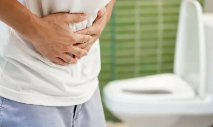 man in white t-shirt holding hands to stomach with toilet in background