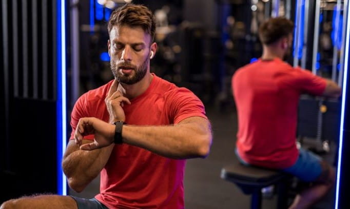 man in a red shirt holding two fingers to his neck and looking at a smartwatch while checking his heart rate in a gym