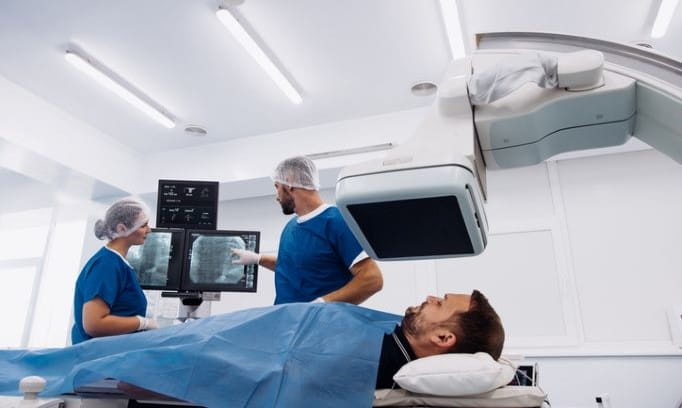 Medical providers discuss a neurointerventional surgery procedure in a modern operating room equipped with advanced imaging technology as a male patient lies on a procedure table.