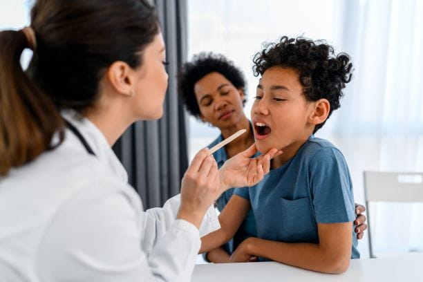 A young boy visiting the doctor with his mom, having his throat checked with a tongue press.