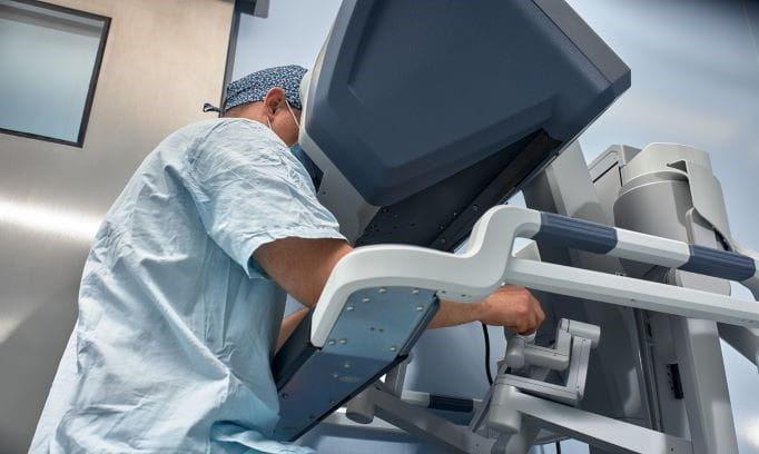 Male surgeon in blue scrubs sitting at a robotic surgical system, operating it with his hands while looking into a large viewing screen.